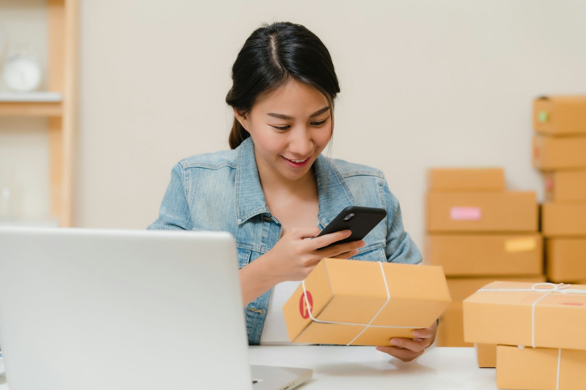 Woman checking a parcel with her phone next to a laptop and boxes